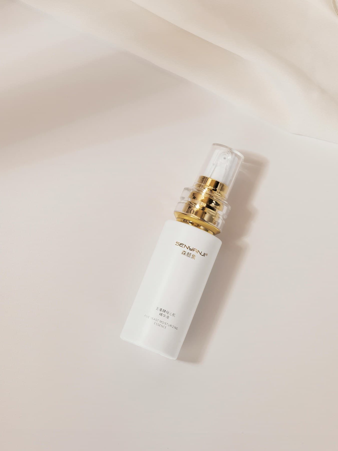 Single serum essence bottle with gold-toned pump on soft ivory backdrop